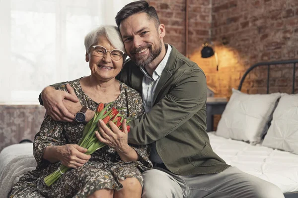 Äldre pensionär dam och hennes tusenåriga son sitter på en säng, kramas och firar mors dag — Stockfoto