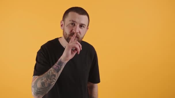 Young Caucasian man wearing a black T-shirt and with a big tatto on his hand putting a finger on mouth and looking into the camera yellow background studio shot medium shot — Stok video