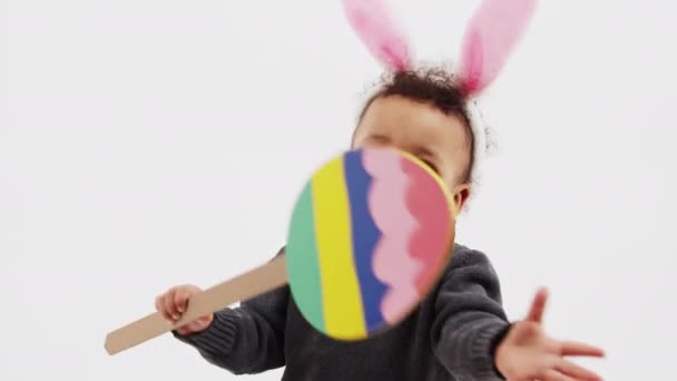 Little African American curly boy with bunny ears walking and holding a big paper Easter egg full shot studio shot white background copy space — Vídeo de stock