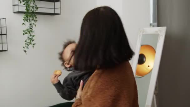 Attraktiv vit mörkhårig kvinna håller en härlig liten pojke med lockigt hår och njuter med honom i det moderna rummet medium shot — Stockvideo