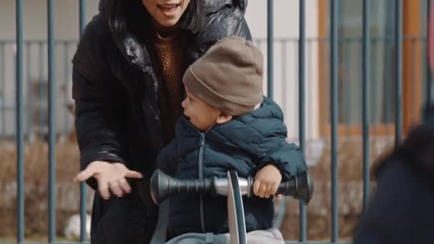 Niño emocionado vistiendo sombrero gris abrigo negro y pantalones grises sentado en una sierra de ver y reír medio tiro al aire libre — Vídeos de Stock