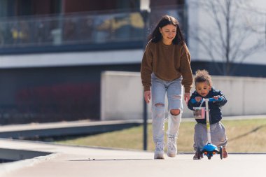 Gülümseyen esmer, beyaz bir anne ilk kez scooter kullanan melez oğlunu destekliyor. Açık hava çekimi. Güneşli hava.