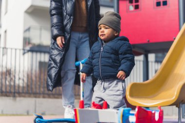 Çocuk parkına bakan, sıcak giysili, tuhaf görünüşlü, melez bir erkek bebek. Bahar zamanı aktiviteleri. Mutlu çocukluk kavramı.