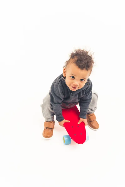 Vertical studio shot of a cute innocent biracial baby boy looking at camera and holding red skateboard over white background. — Photo