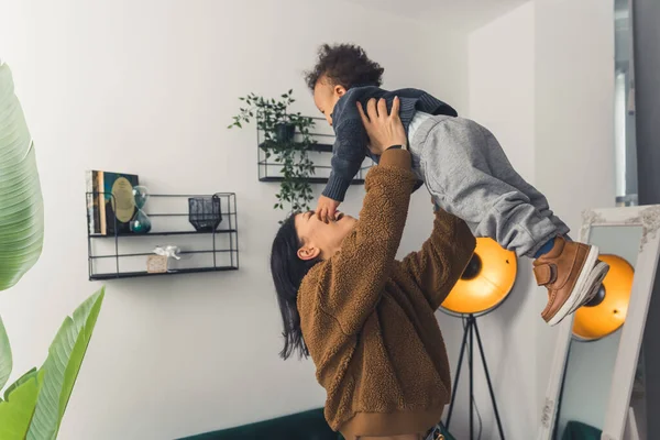 Young caucasian dark-haired mum lifting her biracial adorable son up in their modern flat. Relationship between parent and child. — Stock fotografie
