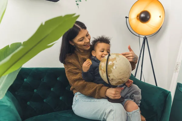 Young beautiful mother sitting on a green couch with her adorable toddler son and teaching him about the world by holding a globe. — Stok fotoğraf