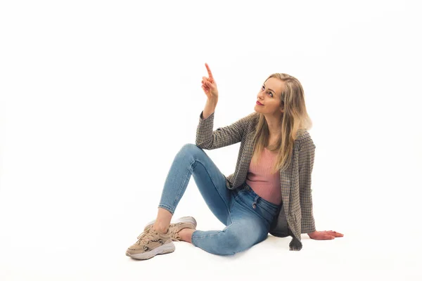 可爱的白人金发姑娘坐在地板上，一个手指指向上方的全镜头演播室拍摄了白色背景的复制空间 — 图库照片