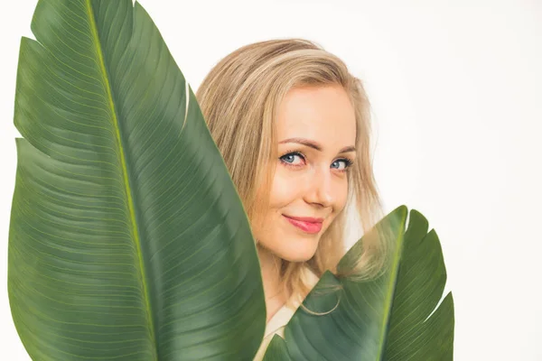 Charming European blue-eyed blonde girl with a pretty smile stands behind the green leaves medium closeup studio shot white background copy space —  Fotos de Stock