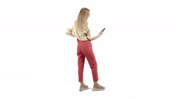 Cute Caucasian blue-eyed blonde girl with pretty smile wearing shirt and pink trousers standing sideways and holding phone studio shot white background medium full shot copy space — Stock video