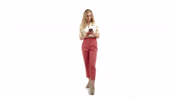 Nice Caucasian blonde girl looking into the phone wearing shirt and pink trousers full shot white background copy space studio shot — Stockvideo