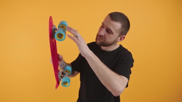 Young satisfied personable bearded European man holding a skateboard orange background studio shot medium shot — стокове відео