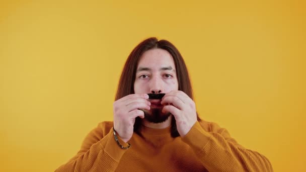 Movember in action. Cheerful caucasian long-haired man wearing yellow sweatshirt and holding fake moustache. — Stockvideo