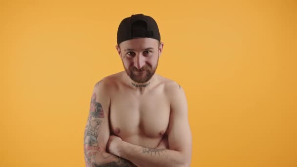 Satisfied shirtless bearded tattoed muscular Caucasian man with cap standing with crossed arms on orange background studio shot indoor medium closeup — Stockvideo