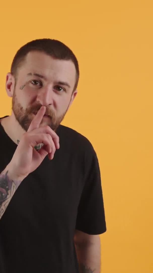 Attractive bearded man with tattoos putting a finger on his mouth orange background studio shot medium shot vertival video — Stockvideo