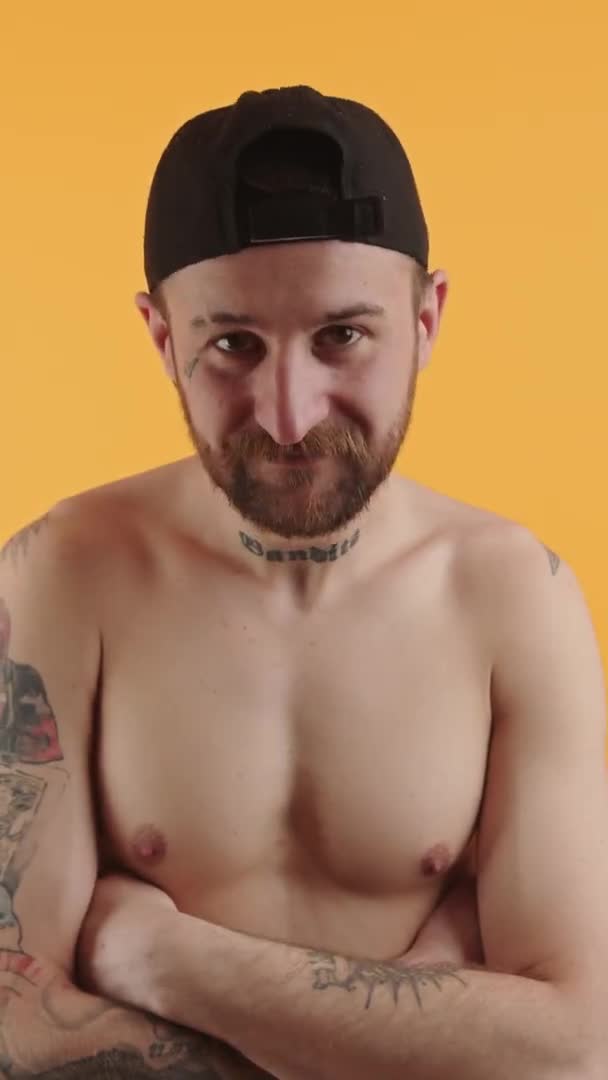 Studio shot of happy shirtless bearded tattoed muscular European man with cap standing with crossed arms orange background indoor medium closeup vertical video — 비디오