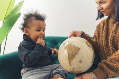 Geography education from a young age concept. Biracial toddler and his caucasian mother sitting on a sofa and looking at a globe.