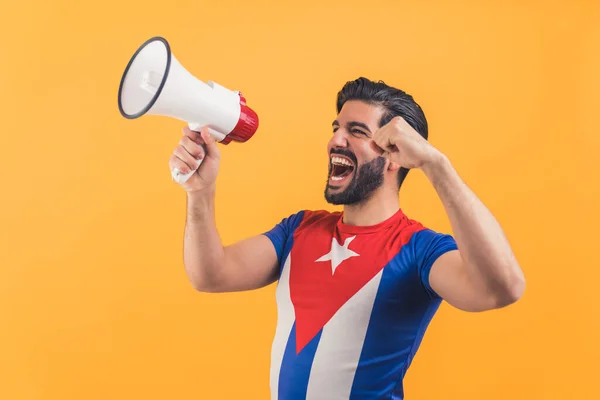 Excited Latin man in the T-shirt of Cuban flag celebrating something and holding a megaphone and shouting orange background medium shot studio shot celebration concept — Stockfoto