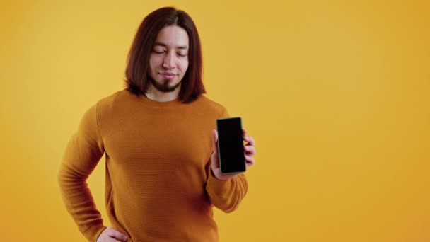 Portrét sebevědomý běloch drží velký smartphone s černou prázdnou obrazovkou v ruce ukazující to na kameru přes žluté pozadí. — Stock video