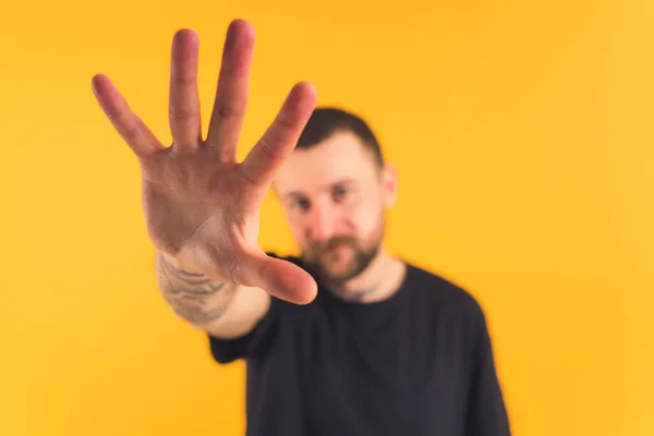 Caucasian bearded tattooed guy stretching his hand to the camera and trying to cover it with his palm, foreground focus. Stop sign. Yellow background. — стоковое фото