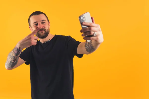 Bearded European guy with tattoo sleeves taking a selfie with his smartphone in studio over yellow background. — ストック写真
