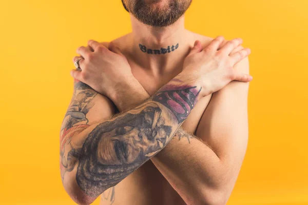 Tattooed shirtless caucasian adult man crosses arms in front of his chest covering his body over yellow background. Body shaming concept. — стоковое фото