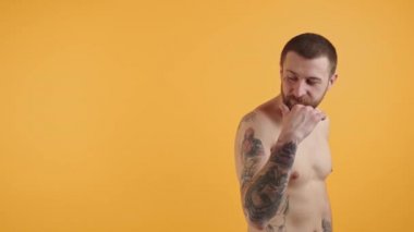 Tişörtsüz, sakallı, beyaz adam arkasına bakıp bir şeyi işaret ediyor. Boşluk sarı arkaplan çekimini kopyala.