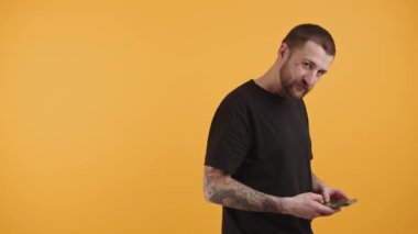 Clever streetwise caucasian guy with tattoo sleeves and a tattoo on his face having a look at camera while scrolling his social media. Studio shot with yellow background.