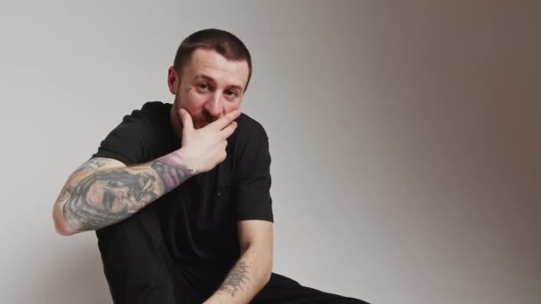 Europeisk skäggig tatuerad kille sitter i en studio över grå bakgrund tittar på kameran och vidrör hans ansiktshår. — Stockvideo