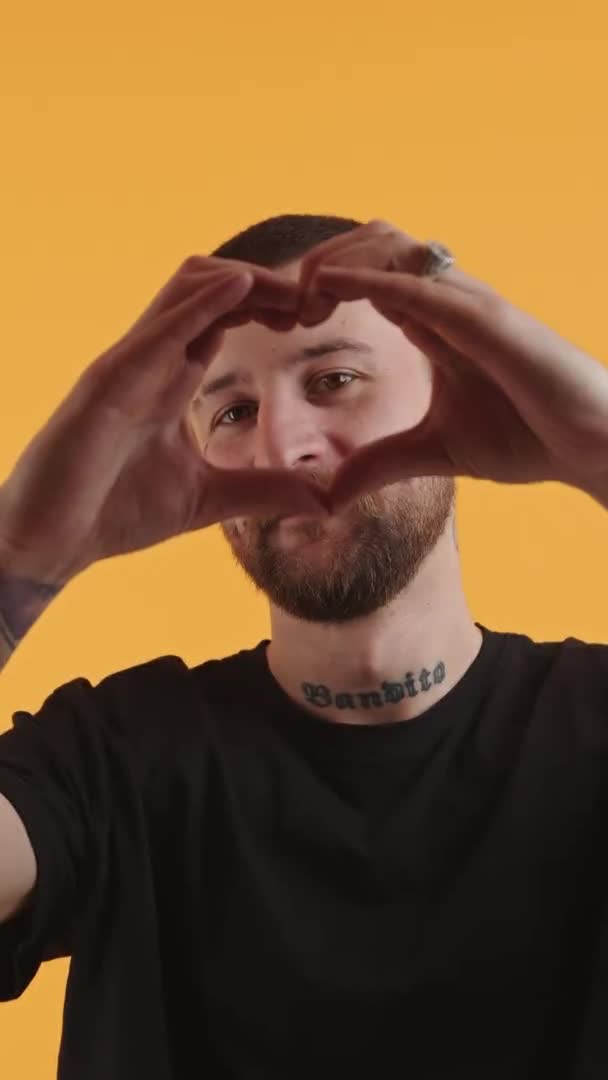 Vertikaler Clip eines kaukasischen Mannes mittleren Alters in einem schwarzen T-Shirt, der eine Geste des Herzens in die Kamera zeigt. Gelber Hintergrund, Studioaufnahme. — Stockvideo