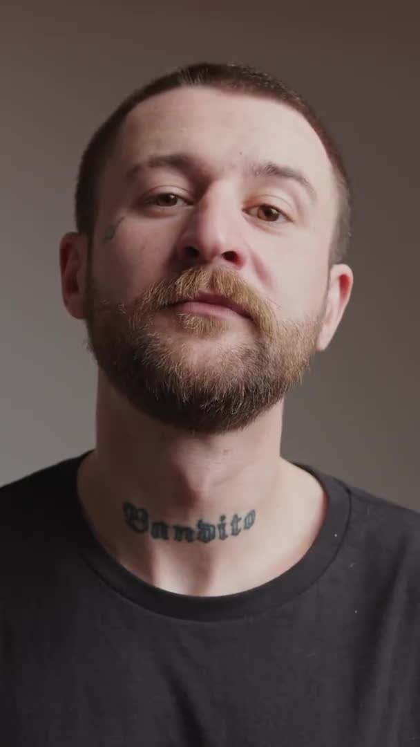 Vertical studio shot of a middle-aged bearded man with tattoo on his neck and chick nodding and looking at camera. — Vídeo de Stock