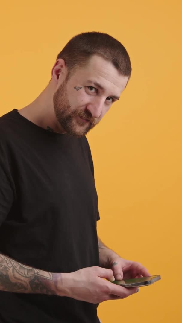 Vertical studio shot of a middle-aged caucasian tattooed guy scrolling social media on his smartphone and looking at camera. Yellow background. — стоковое видео