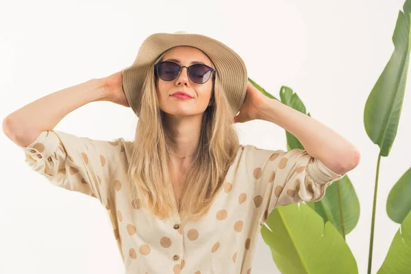 Fascinating Caucasian blonde girl with sunglasses and hands on her beige hat standing in front of camera medium closeup white background with green leaves studio shot copy space — Stock Photo, Image