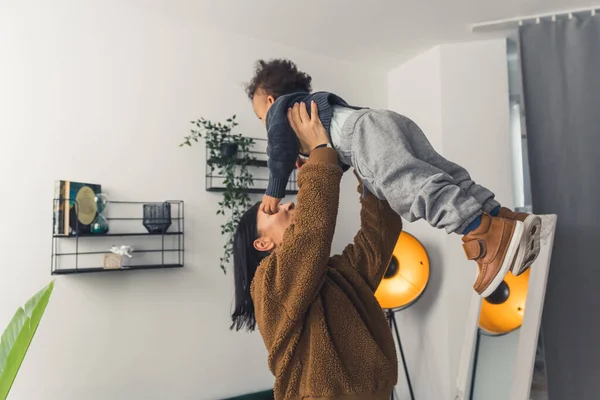 Black haired woman with a brown sweater holding curled little boy upwards in the room with white rooms medium shot — Stock fotografie