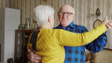 Oturma odasında gülümseyen ve dans eden yaşlı adam ve kadının orta boy fotoğrafı.