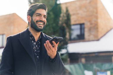 Siyah elbiseli, genç ve yakışıklı bir Latin Amerikalı bir adam elinde bir telefon tutuyor ve açık hava kentsel arka planında gülümsüyor. 