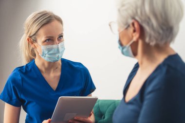 Zeki, kendine güvenen kadın sağlık çalışanı. Elinde bir pano ya da tablet tutarken yaşlı emeklileri muayene ediyor..