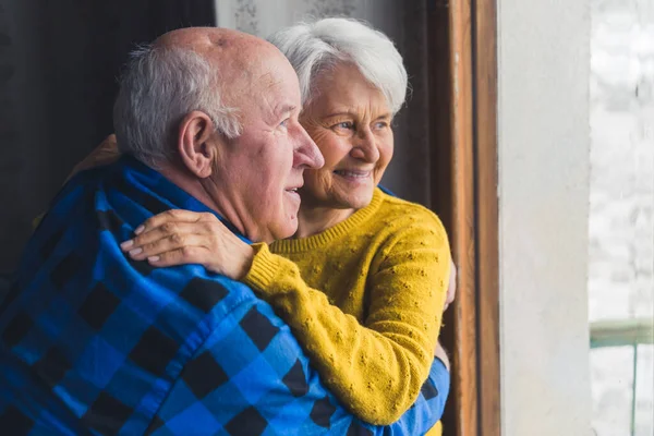 Indoor portrait of a happy caucasian heterosexual retired couple of seniors standing by the window embracing each other. — ストック写真