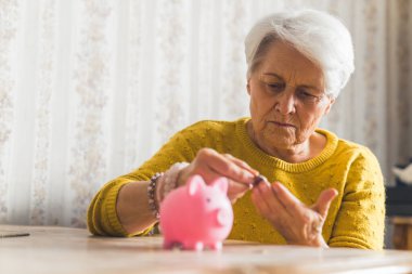 Investments and savings concept. Poor caucasian pensioner gray-haired lady in yellow sweater looks at her money and puts coins to her pink piggy bank.