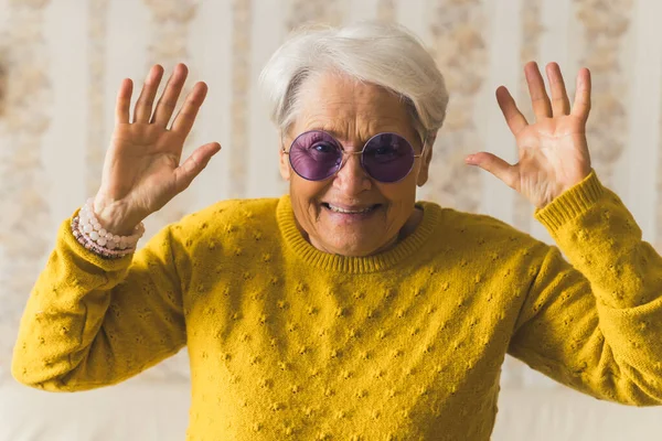 Funny caucasian grandmother in purple sunglasses looking at camera and smiling, and holding her hands up. Vintage wallpaper in the background. — стоковое фото