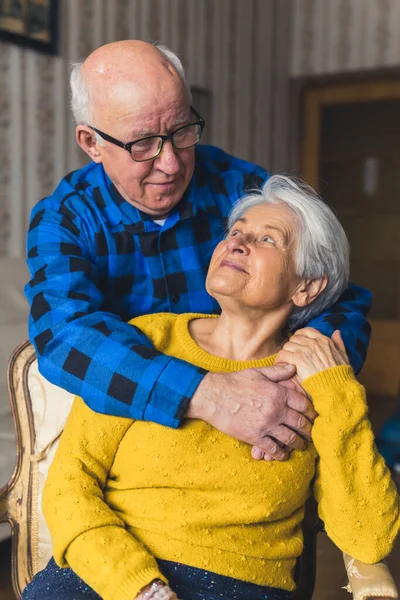 Vertical shot of a hugging elderly caucasian couple. Senior gray-haired woman sitting on an old-fashioned chair being embraced by her bald charming husband in a flannel shirt. — Stockfoto