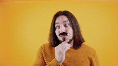 Kımılda. Sarı kazak giyen ve sarı arka planda sahte bıyık takan neşeli, beyaz saçlı bir adam..