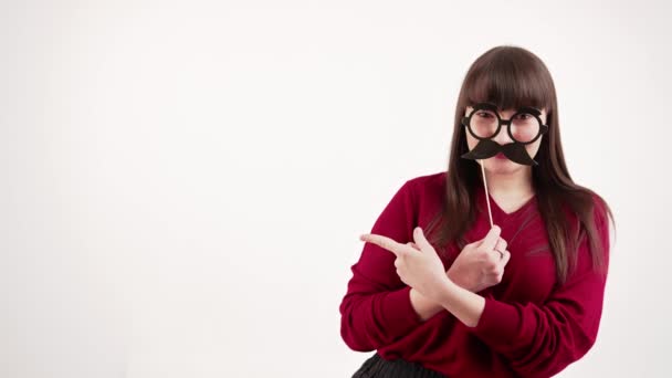 Copy space studio shot of a young caucasian brunette shushing while holding fake mustache and glasses set in front of her face. — ストック動画
