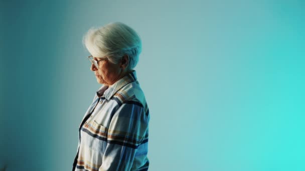 Senior European woman with white hair and glasses standing sideways and thinking about a disease she has copy space blue background medium closeup healthcare concept — стоковое видео