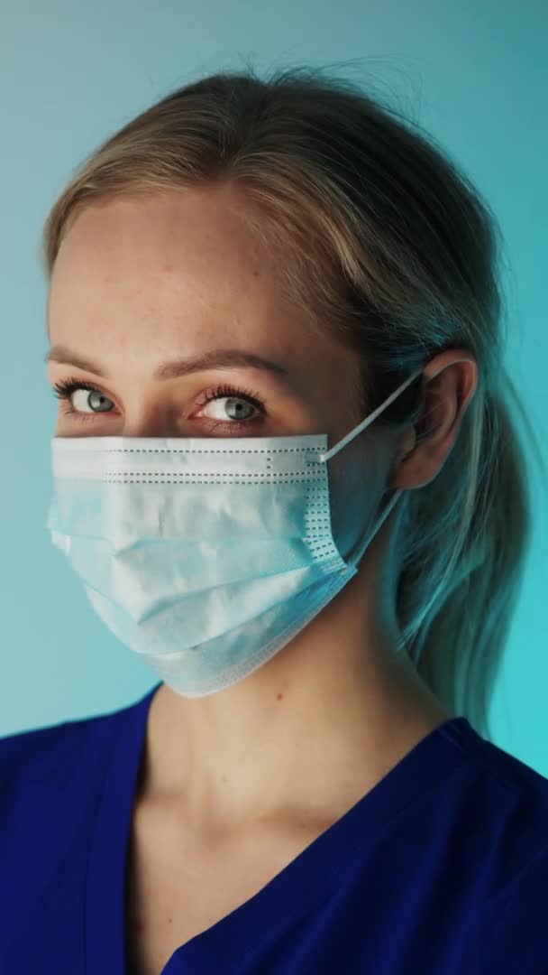 Blond vit sjuksköterska med en kirurgisk mask tittar på kameran vertikal video närbild studio sköt svart bakgrund medicinskt koncept — Stockvideo