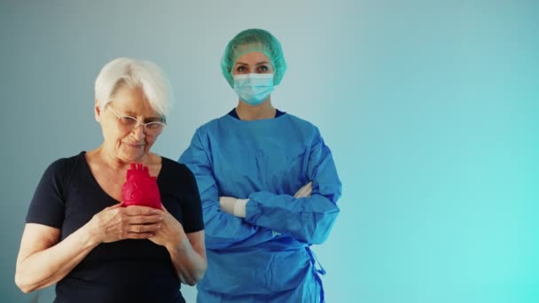Senior femme caucasienne tenant un cœur artificiel et debout devant un médecin bleu fond moyen plan studio concept de soins de santé — Video