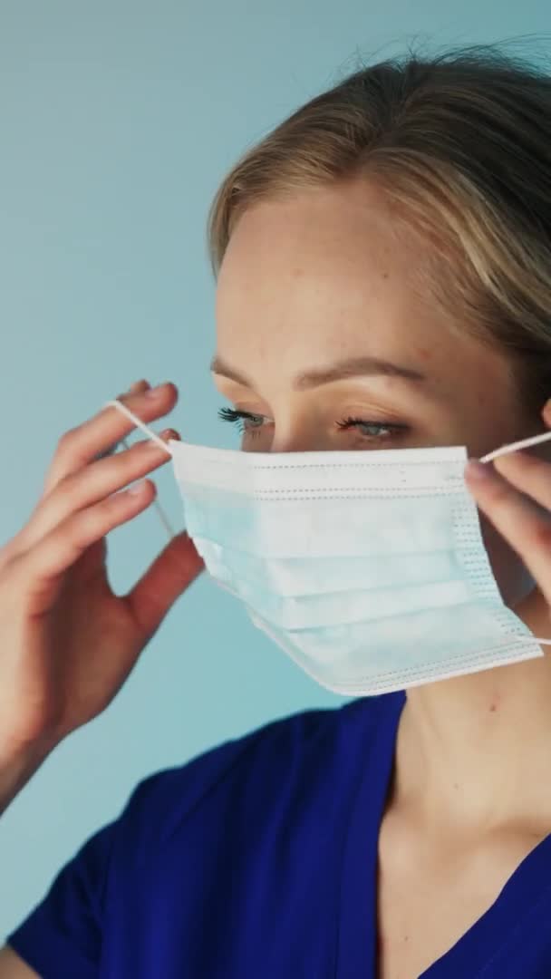 Perawat pirang Eropa menyesuaikan masker bedah pada wajah closeup vertikal studio video menembak biru latar belakang konsep medis — Stok Video