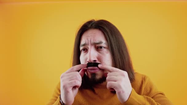 Funny caucasian long-haired bearded guy in yellow sweater touching his black fake moustache. Movember concept. Yellow background studio shot. — Vídeos de Stock