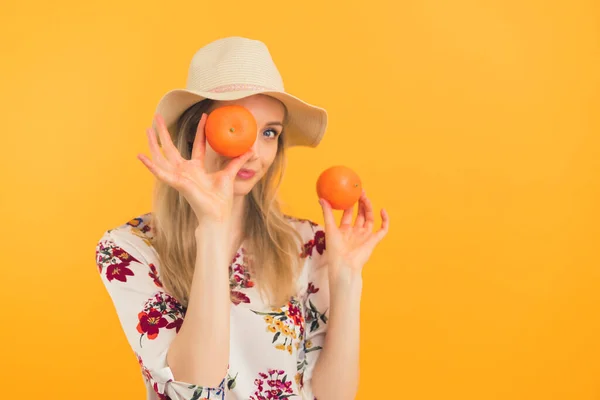 Youthful caucasian blonde girl in her 20s in a hat posing with two oranges or mandarins. Medium studio shot over orange background. — Foto de Stock