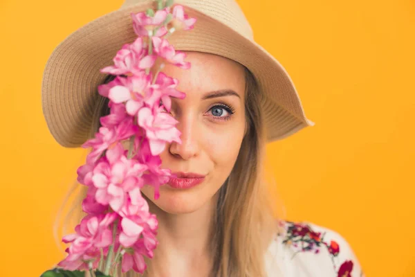 Closeup portrait of adorable caucasian blonde woman in a hat holding beautiful flour in front of her face and covering her eye over orange background. — Stock Photo, Image