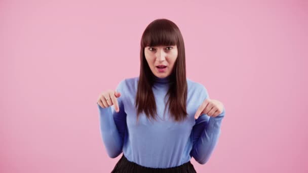 Smiley Europeiska brunett kvinna bär violett polotröja pekar på kopia utrymme under henne över rosa bakgrund. Medium studio skott. — Stockvideo